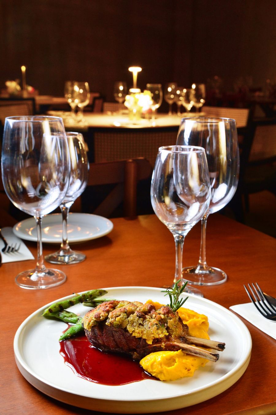 Prato da Toscana Trattoria: carr\u00e9 de cordeiro com crosta de ervas, aspargos e pur\u00ea, em mesa posta \u00e0 luz de velas.