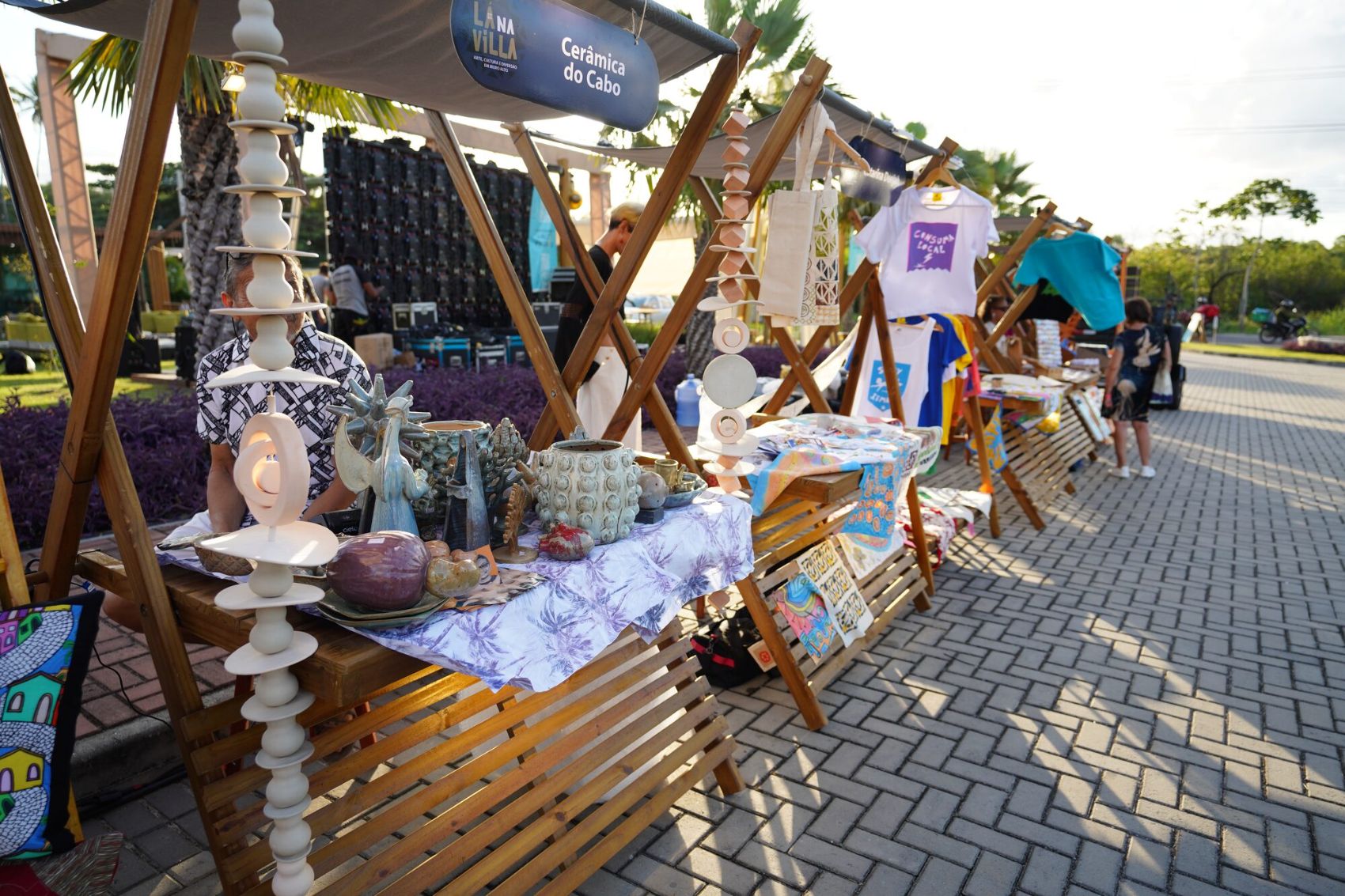 Feira criativa da Villa Muro Alto ao entardecer: barracas de madeira com cer\u00e2mica do Cabo, camisetas e pe\u00e7as autorais de artes\u00e3os da regi\u00e3o.