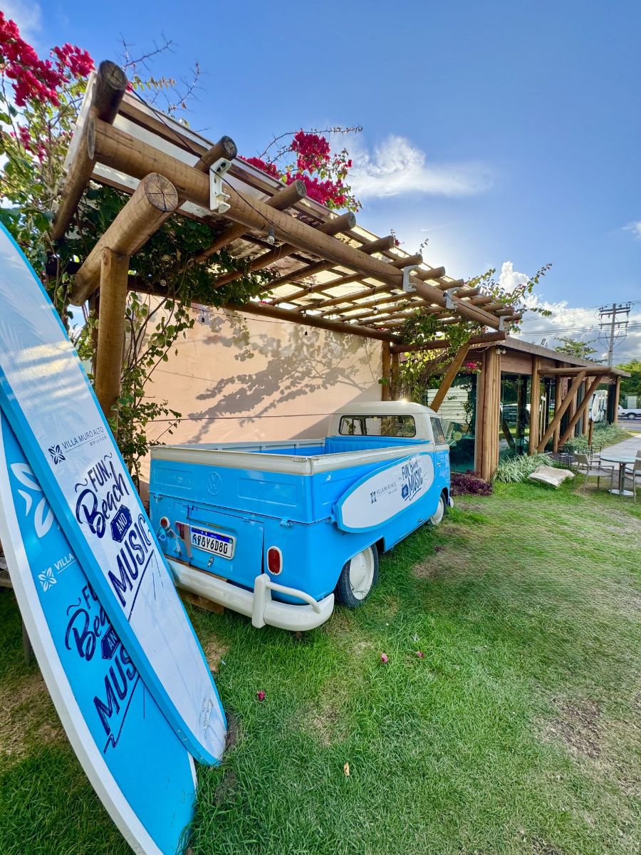 Kombi azul da Villa Muro Alto sob a pergolada florida, com pranchas pintadas com Fun, Beach and Music.