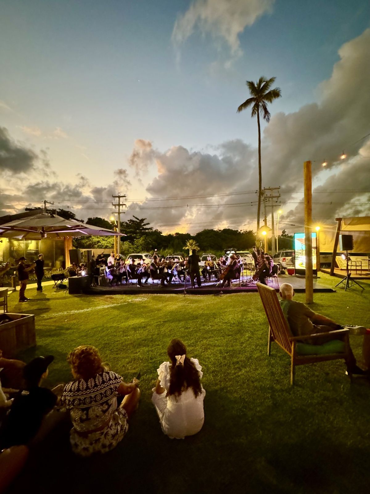 Concerto de orquestra ao entardecer no gramado da Villa, com p\u00fablico sentado na grama e nas cadeiras de madeira.